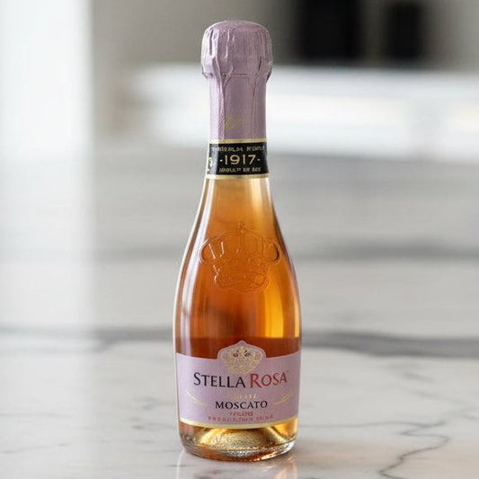 A bottle of Stella Rosa Moscato sparkling wine with a pink label and silver foil, standing on a light wood tabletop against a plain off-white wall.