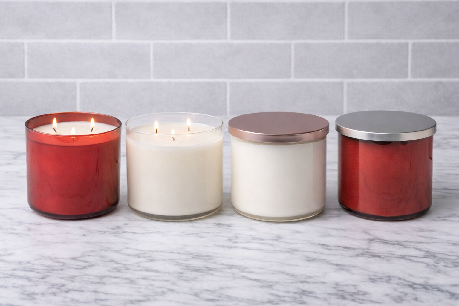 Four candles with different colored glass and metal lids on a marble surface with a tiled wall background.