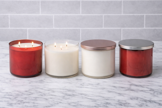 Four candles with different colored glass and metal lids on a marble surface with a tiled wall background.